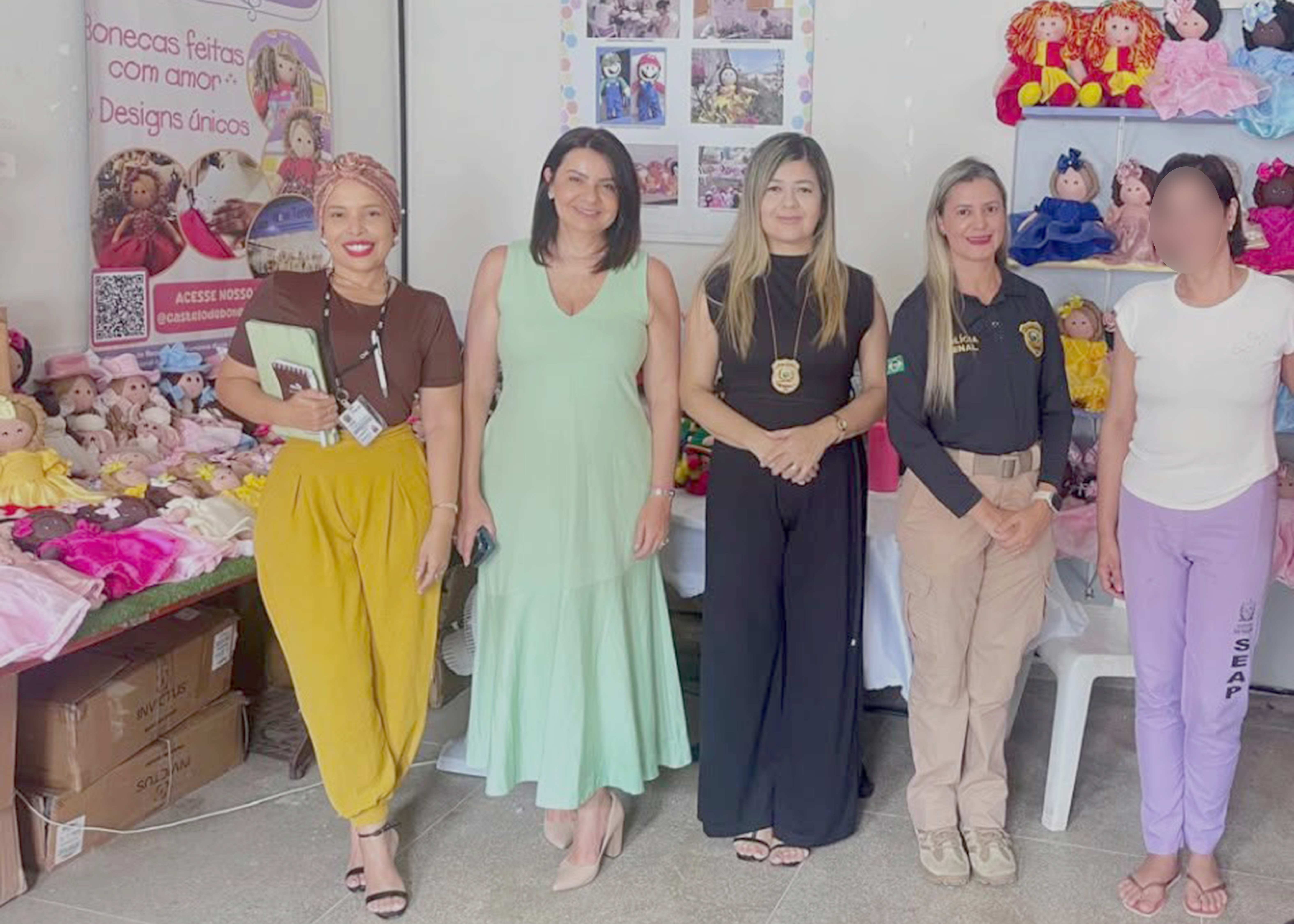 Fotografia em ambiente interno mostrando cinco mulheres lado a lado, em frente a uma exposição de bonecas artesanais de pano. À esquerda, uma mesa e um banner com fotos das bonecas e a frase "Bonecas feitas com amor". Ao centro e à direita, prateleiras exibem bonecas coloridas.  As quatro primeiras mulheres sorriem. A primeira usa blusa marrom, calça amarela e turbante rosa, segurando uma pasta e um caderno. A segunda veste um vestido longo verde-claro. A terceira usa conjunto preto e um distintivo pendurado no pescoço. A quarta veste uniforme preto e bege com distintivo. A quinta pessoa, à direita, está com o rosto desfocado, usa camiseta branca, calça lilás com a inscrição "SEAP" e chinelos. O ambiente sugere uma visita institucional a um ateliê de artesanato.