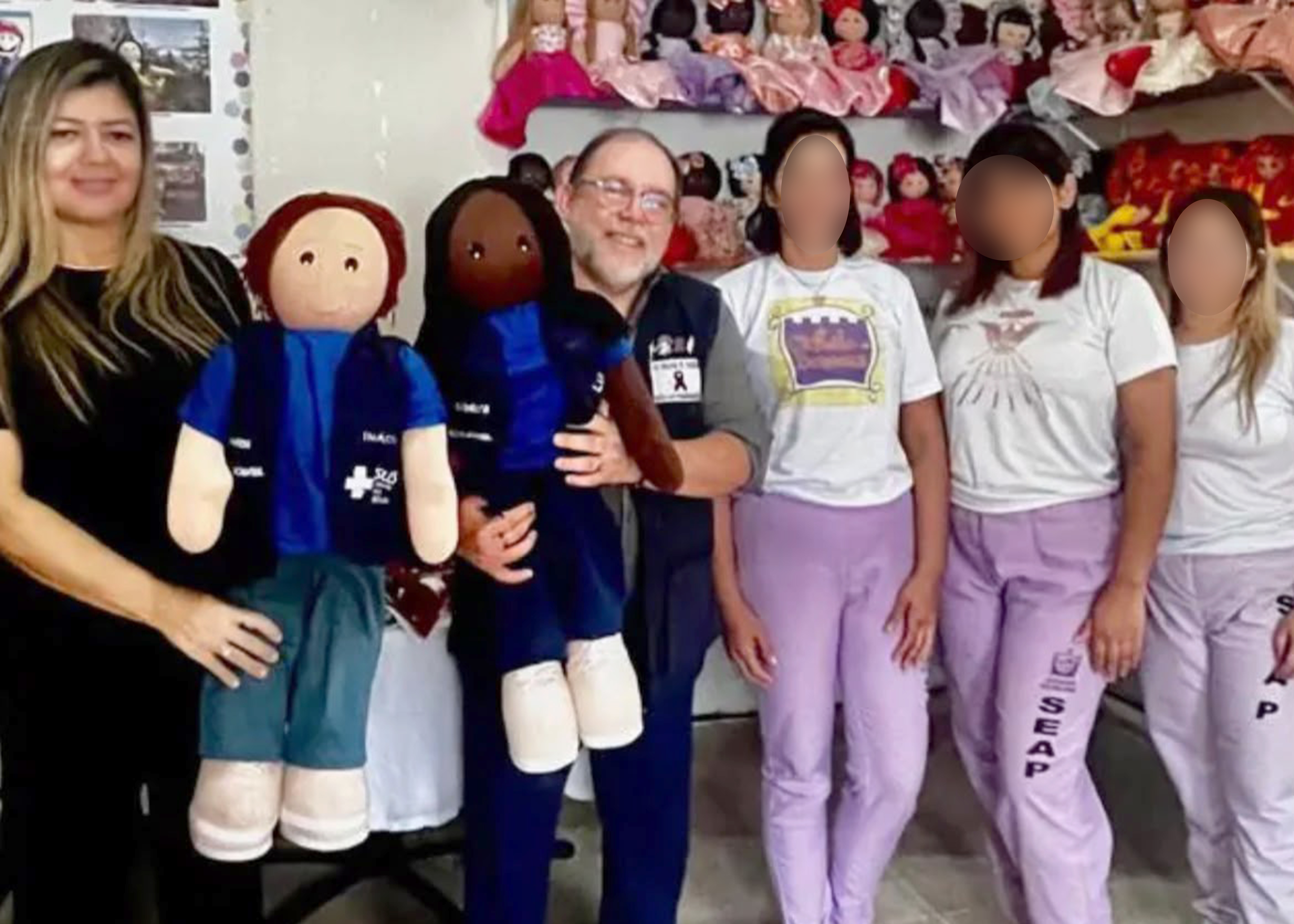 Fotografia em ambiente interno mostrando seis pessoas em um ateliê de bonecas artesanais. À esquerda, uma mulher veste roupa preta e segura uma boneca de pano. Ao centro, um homem de óculos, com colete azul, sorri enquanto segura outra boneca. À direita, quatro mulheres com os rostos desfocados usam camisetas brancas e calças lilás com a inscrição "SEAP". Ao fundo, prateleiras exibem diversas bonecas coloridas, criando um ambiente de artesanato e exposição.